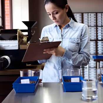 Femme tenant un presse-papiers et notant des observations, avec une machine à torréfier le café en arrière-plan, lors d'une dégustation de café