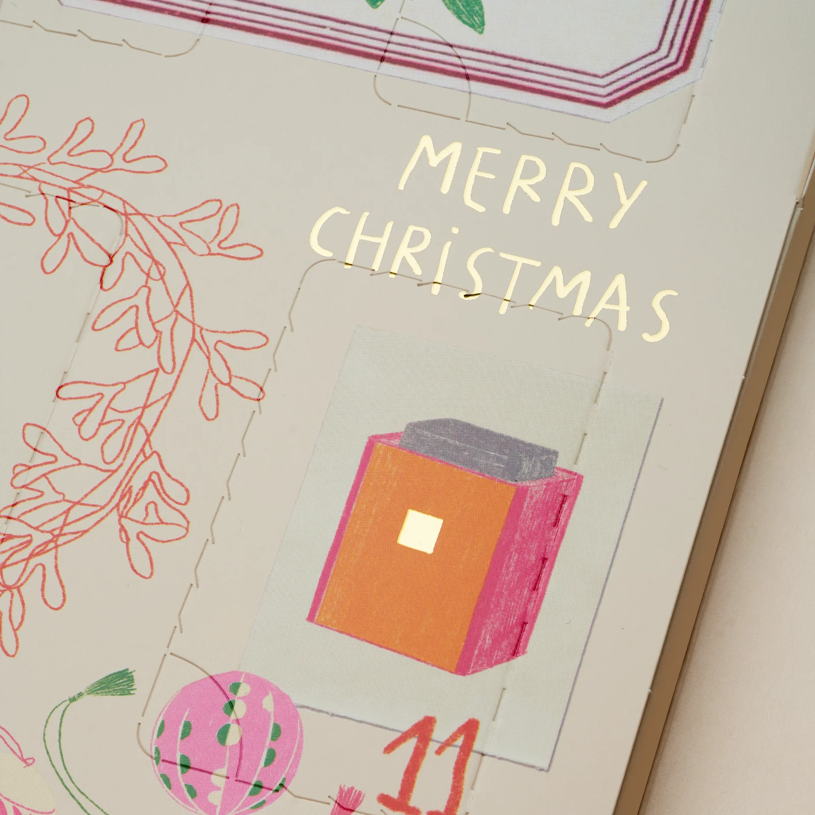 Calendrier de l'avent illustré avec le texte doré Merry Christmas, mettant en avant une case numéro onze représentant un cadeau orange et rose