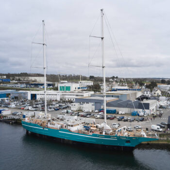 Bateau à voile turquoise avec deux mâts dans un port industriel, nom 'ANEMOS' visible sur la coque