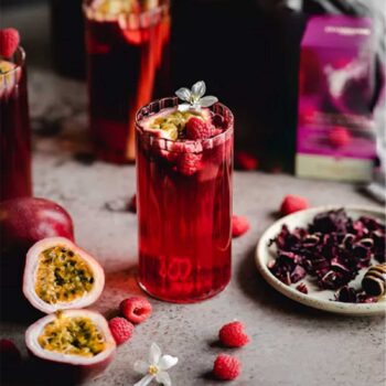 Verre de boisson rouge vif garni de framboises et de fruits de la passion, avec des fruits frais et des fleurs sur une table, évoquant l'infusion glacée Passion Framboise Dammann Frères