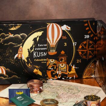 Calendrier de l'Avent Kusmi Tea ouvert, dévoilant des illustrations de voyage avec une fusée, un ballon, et des personnages sur un éléphant, devant une carte ancienne et des boîtes de thé.