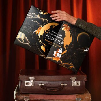 Calendrier de l'Avent Kusmi Tea orné d'une illustration cosmique, avec un globe terrestre, une fusée et un voilier, posé sur des valises vintage.