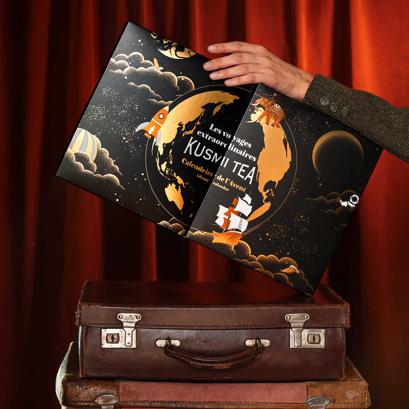 Calendrier de l'Avent Kusmi Tea orné d'une illustration cosmique, avec un globe terrestre, une fusée et un voilier, posé sur des valises vintage.