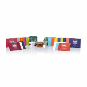 Présentation complète du contenu du Calendrier de l’Avent Kusmi Tea 2024 avec les miniatures de thé, un pot de spécialité à tartiner, un infuseur étoile et plusieurs sachets de thés colorés. Les sachets sont disposés en arc autour des autres éléments.