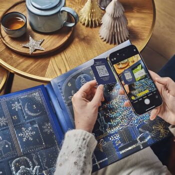 Une main tient un petit carton bleu avec un QR code devant un calendrier de l'Avent ouvert, tandis qu'un smartphone capture l'image du QR code