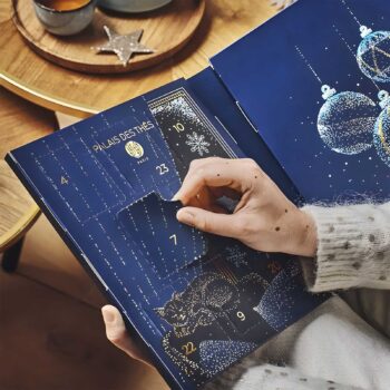 Main d'une personne ouvrant une case numérotée sur un calendrier de l'Avent bleu nuit orné de motifs dorés, révélant une sélection de thés Palais des Thés