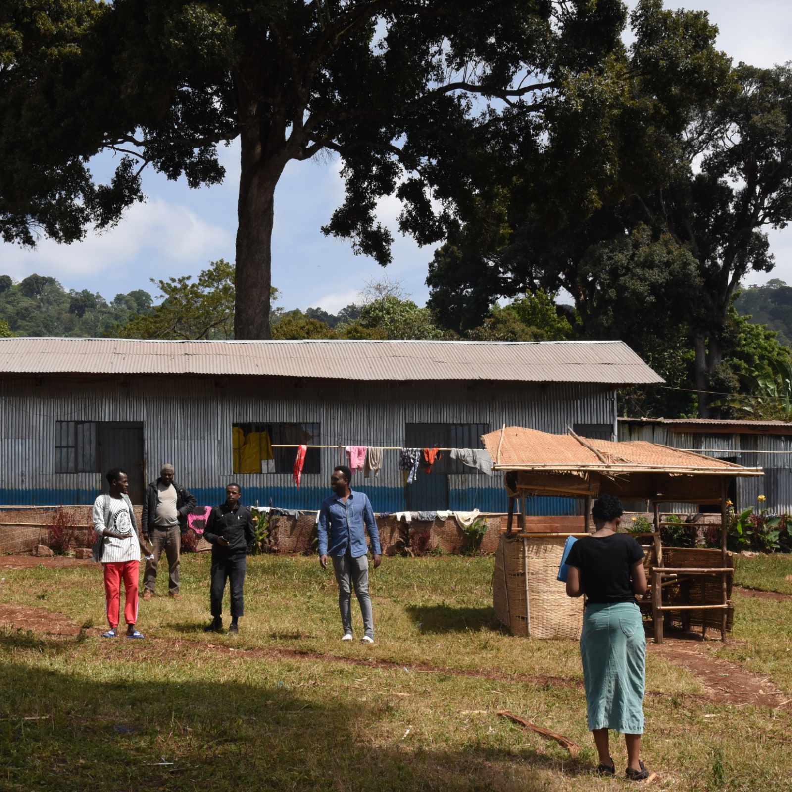Des personnes se tiennent dans un espace extérieur devant un bâtiment en tôle ondulée, avec du linge séchant sur une corde. Un grand arbre domine l'arrière-plan.