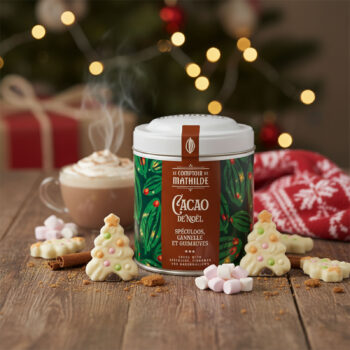 Boîte de cacao de Noël du Comptoir de Mathilde entourée de biscuits en forme de sapin, guimauves et bâton de cannelle, avec une tasse de chocolat chaud fumant en arrière-plan