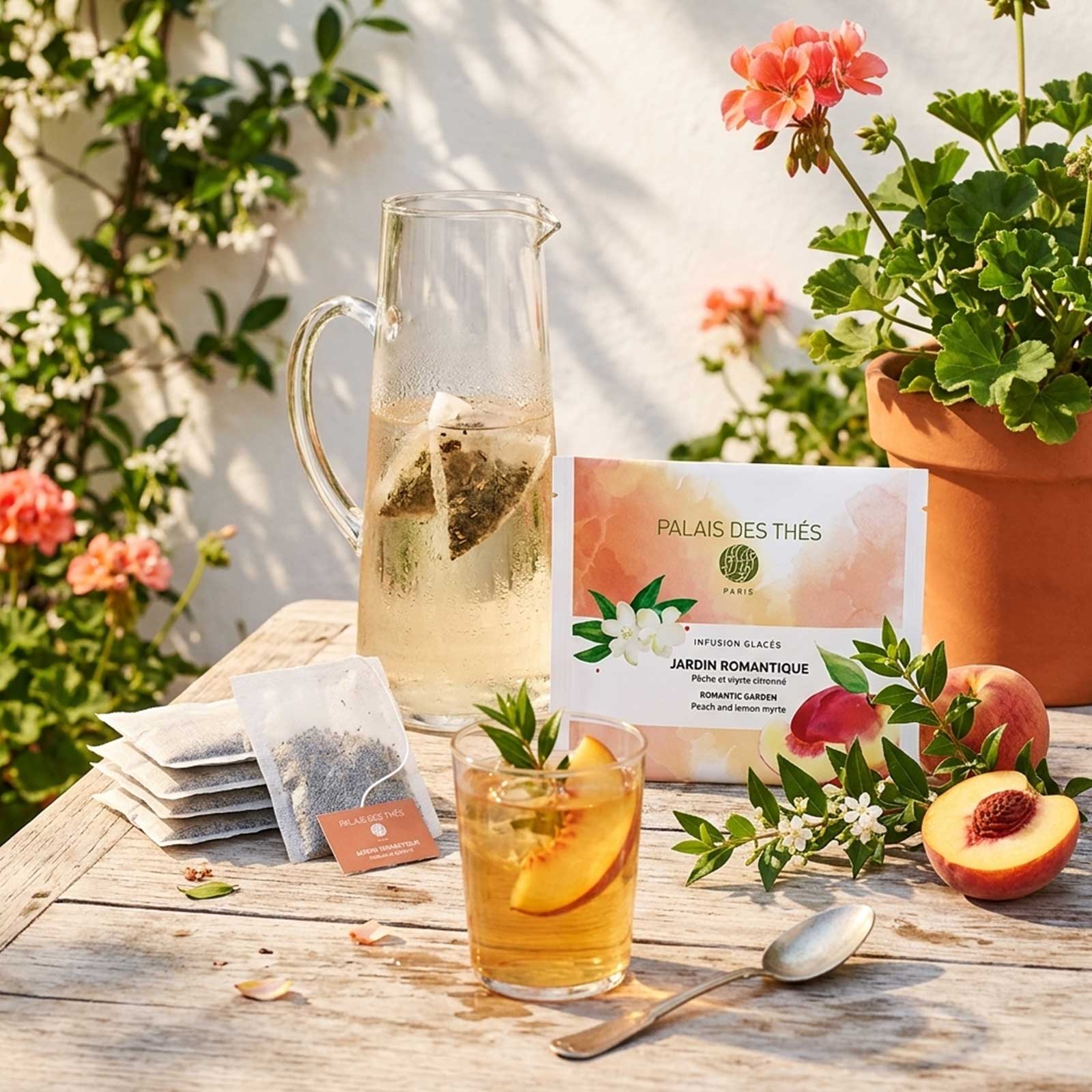 Carafe en verre remplie d'eau et de sachets de thé glacé Jardin Romantique, avec un verre de thé glacé garni de pêche et une boîte de Palais des Thés à côté