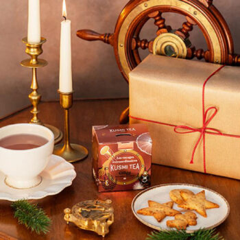 Boîte de thé Kusmi Tea édition Glögg 2024 posée sur une table en bois, entourée de bougies, d'un gouvernail, d'un cadeau emballé et de biscuits en forme d'étoiles.