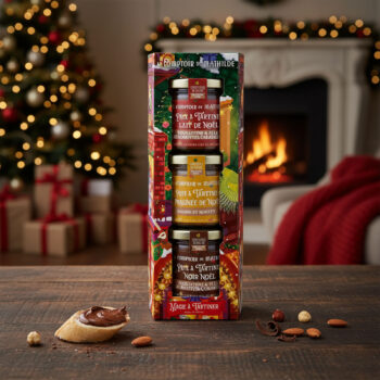 Coffret cadeau Le Comptoir de Mathilde avec trois pots de pâtes à tartiner de Noël sur une table en bois, une tranche de pain tartinée de chocolat à proximité