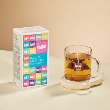 Boîte d'assortiment Kusmi Tea Les Bien-être avec ses carrés colorés et un mug en verre contenant une infusion sur un socle en pierre