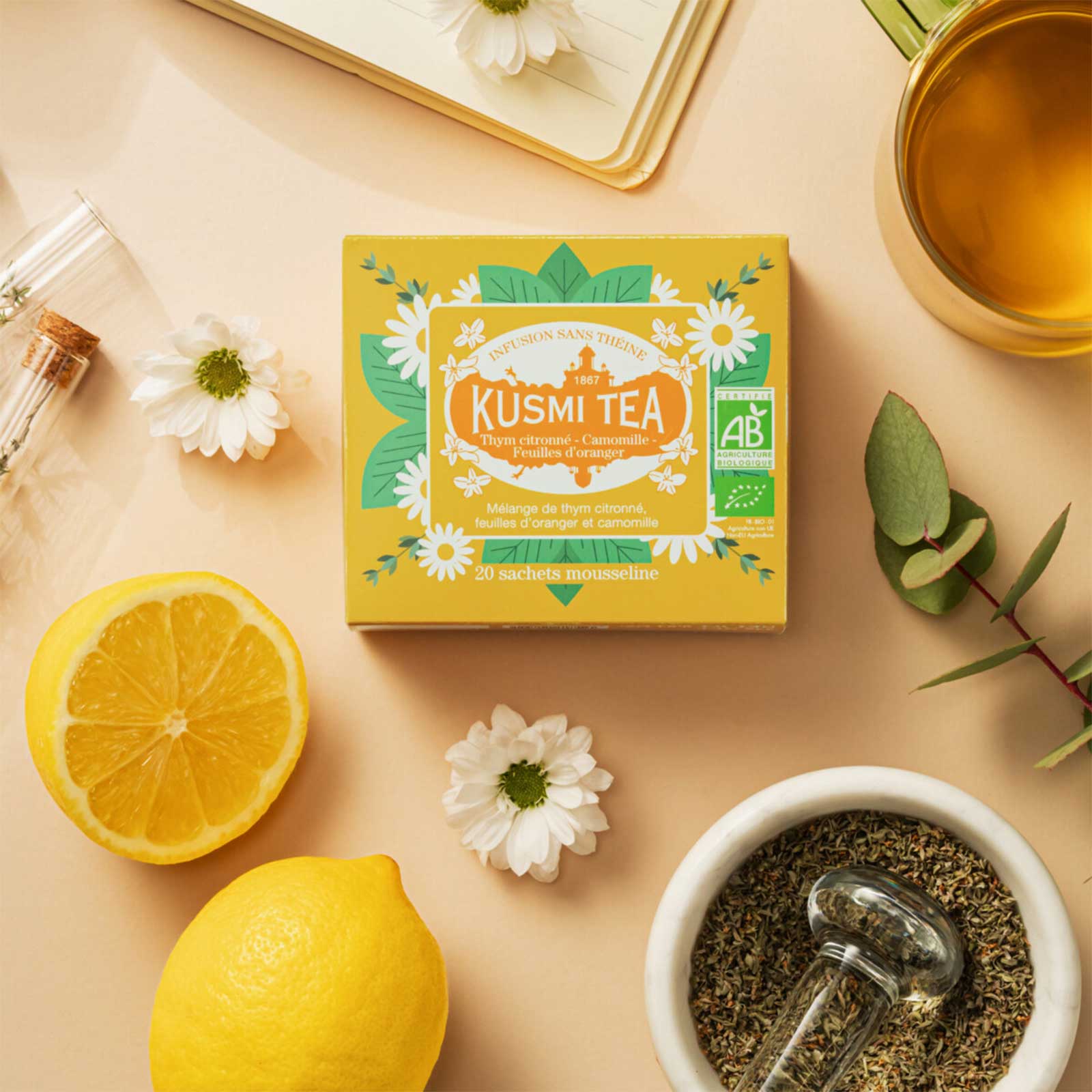 Boîte de Kusmi Tea infusion sans théine thym camomille feuilles d'orange bio, entourée de citrons, fleurs et d'une tasse de thé.