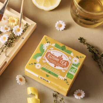 Boîte jaune de Kusmi Tea infusion sans théine aux herbes, avec des motifs de fleurs de camomille et des feuilles vertes, mentionnant thym citronné, camomille et feuilles d'oranger.