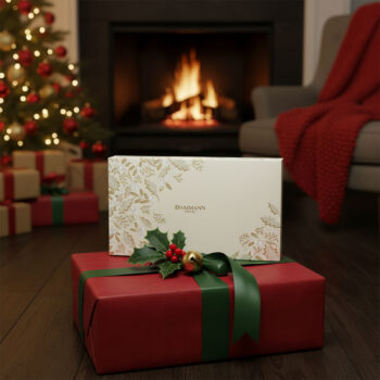 Coffret Dammann Frères Rêves de Noël rouge et or, décoré de motifs végétaux, posé sur un paquet cadeau festif près d'une cheminée et d'un sapin de Noël