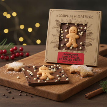 Tablette de chocolat au lait du Comptoir de Mathilde avec un bonhomme en pain d'épices, perles croustillantes blanches et chocolatées, sur une planche en bois.