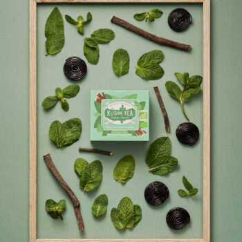 Boîte de thé Kusmi Tea Réglisse-Menthe entourée de feuilles de menthe fraîche, bâtons de réglisse et bonbons de réglisse, le tout sur fond vert pastel