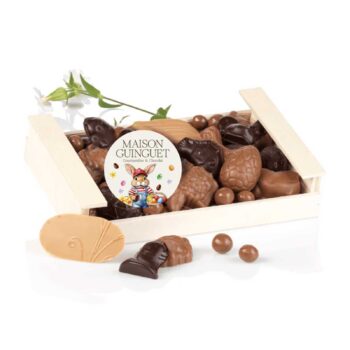 Cagette en bois MAISON GUINGUET remplie de chocolats de Pâques variés, dont des lapins et des œufs, avec un autocollant festif