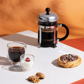 Cafetière Bodum Chambord inox remplie de café, servie avec deux verres, une petite coupelle de crème et une pâtisserie aux noix