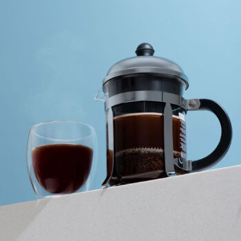 Cafetière à piston Bodum Chambord en inox avec un verre de café noir fumant à côté, mise en scène sur un plan de travail blanc