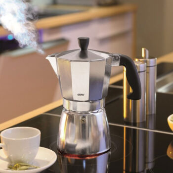 Cafetière italienne Gefu Lucino en aluminium poli sur une plaque de cuisson à induction, dégageant de la vapeur, avec une tasse à café à côté