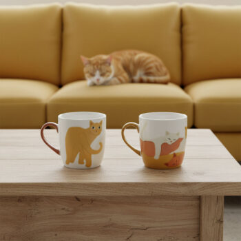 Deux mugs en céramique blancs décorés de motifs de chats, posés sur une table en bois. Un chat roux est visible flou en arrière-plan sur un canapé jaune.