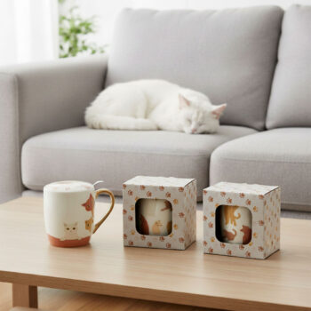 Tisanière Gustave Amadeus en porcelaine avec motifs de chats, accompagnée de sa boîte, sur une table en bois, avec un chat blanc dormant sur un canapé en arrière-plan.