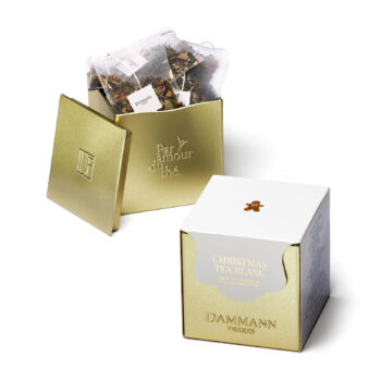 Boîte métallique dorée ouverte, remplie de sachets de thé Dammann Frères, à côté d'une boîte cadeau blanche ornée d'un petit bonhomme en pain d'épices, étiquetée 'Christmas Tea Blanc'