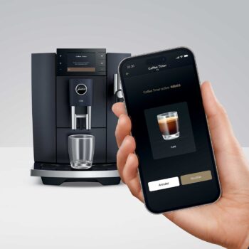 Main tenant un smartphone affichant l'application J.O.E. de JURA, avec une machine à café JURA E8 en arrière-plan. La fonction Coffee Timer est active.