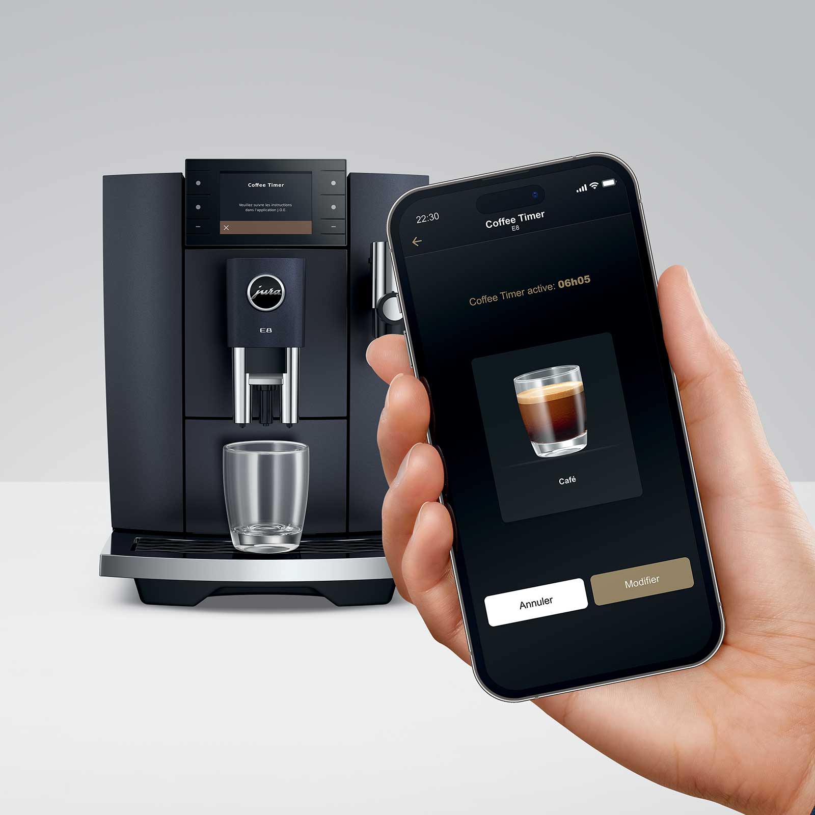 Main tenant un smartphone affichant l'application J.O.E. de JURA, avec une machine à café JURA E8 en arrière-plan. La fonction Coffee Timer est active.
