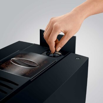 Main ajustant le bouton de réglage de la finesse de mouture sur une machine à café JURA E8, avec un réservoir de grains de café visible