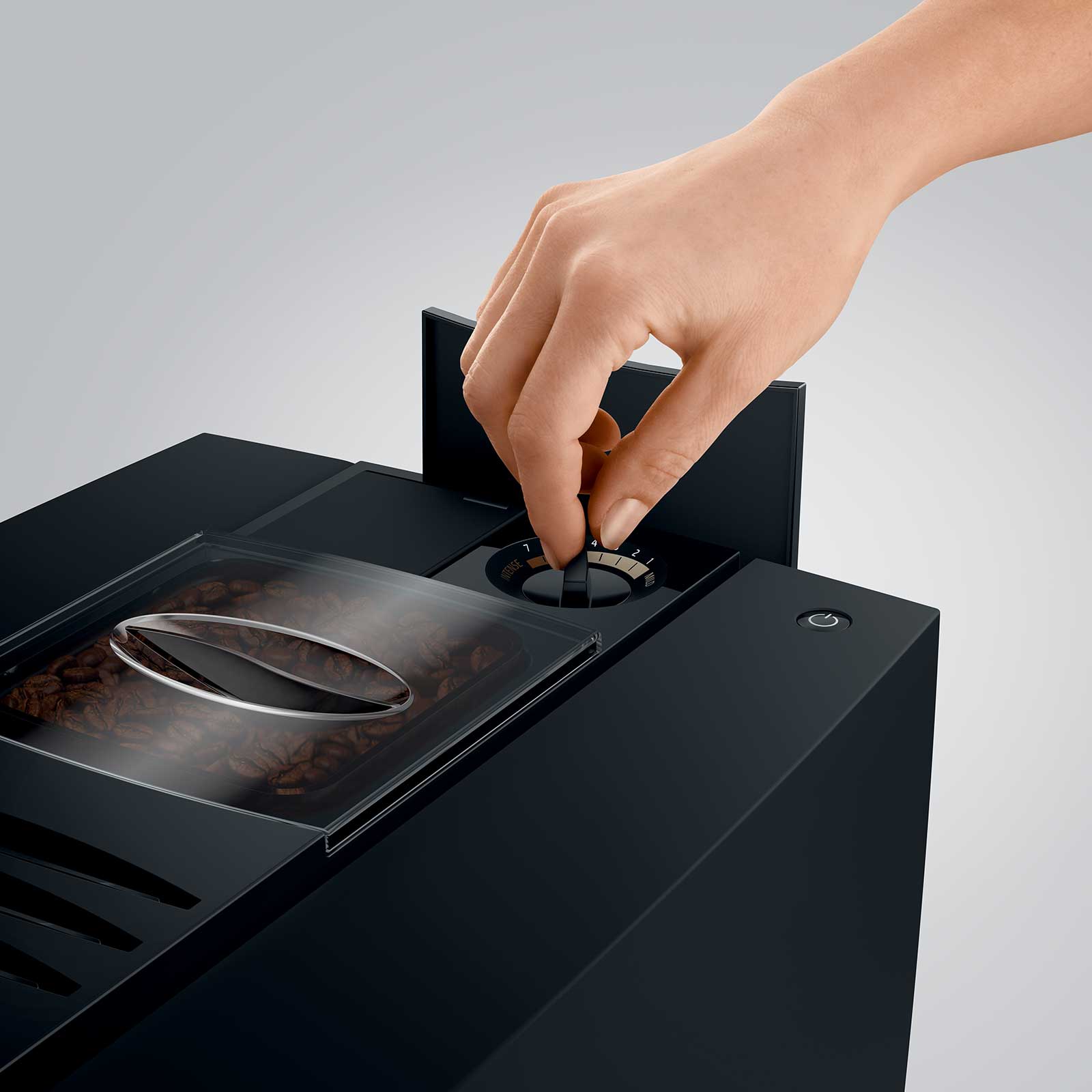 Main ajustant le bouton de réglage de la finesse de mouture sur une machine à café JURA E8, avec un réservoir de grains de café visible