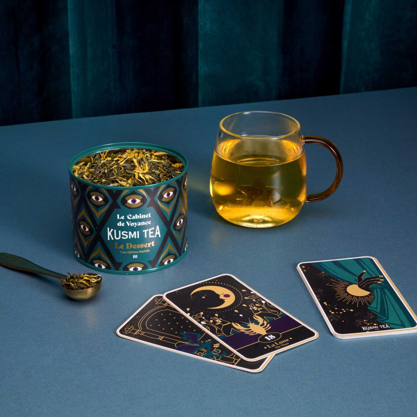 Boîte de thé Kusmi Tea Le Cabinet de Voyance posée à côté d'une tasse en verre remplie d'infusion et de cartes de tarot sur un fond bleu foncé