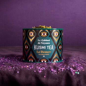 Boîte de thé Kusmi Tea Le Cabinet de Voyance sur un fond violet, décorée d'un motif d'yeux dorés et entourée de paillettes violettes