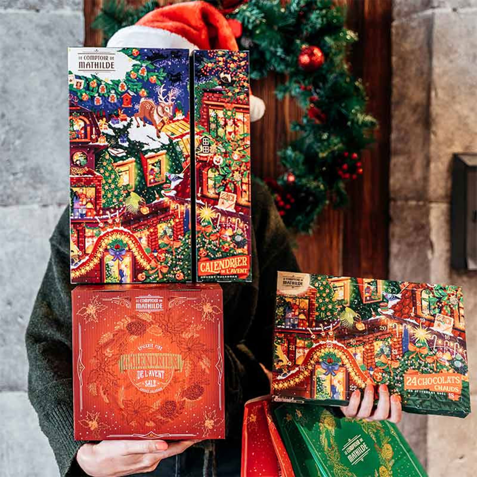 Personne tenant des calendriers de l'avent festifs du Comptoir de Mathilde remplis de chocolats chauds, décorations de Noël en arrière-plan