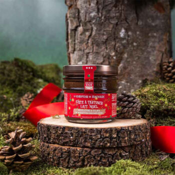 Bocal de pâte à tartiner Lait Noël du Comptoir de Mathilde, sur une souche en bois décorée de mousse et de pommes de pin, avec un ruban rouge.