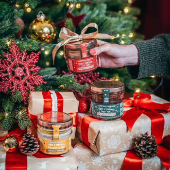 Trois pots de pâte à tartiner Comptoir de Mathilde décorés pour Noël, dont une tenue à la main, entourés de cadeaux et de décorations de sapin.