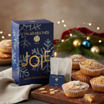 Boîte de thé Palais des Thés "Joie" bleue nuit avec flocons dorés, entourée de petits gâteaux aux amandes et décorations de Noël lumineuses.