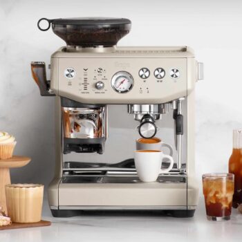 Machine à expresso Sage The Barista Express Impress Nougat avec moulin intégré, préparant un expresso dans une tasse blanche, entourée de quelques pâtisseries et d'une boisson glacée.