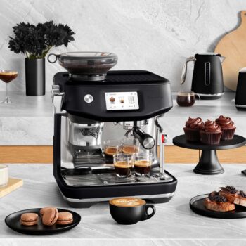 Machine à expresso Sage The Barista Touch Impress noire avec moulin intégré, produisant des espressos dans des tasses, entourée de pâtisseries et d'une bouilloire.