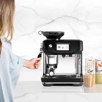 Femme utilisant la machine à expresso Sage Barista Touch Impress noire, avec des briques de lait d'avoine et d'amande en arrière-plan