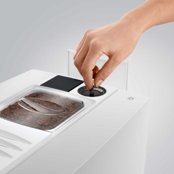 Main ajustant le bouton de réglage de la mouture sur une machine à café JURA E8 Piano White, avec des grains de café visibles dans le réservoir