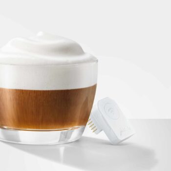 Verre de café latte avec mousse onctueuse à côté d'un adaptateur Jura Wifi Connect v2 blanc, illustrant la connectivité des machines à café