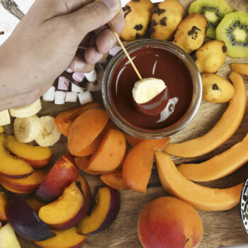 Une main trempe une tranche de banane dans un pot de fondue au chocolat au lait, entouré de fruits coupés, guimauves et biscuits