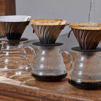 Trois cafetières Hario V60 Dripper en métal avec filtres en papier, l'une recevant un filet d'eau chaude pour l'extraction du café