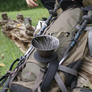 Hario V60 Dripper en métal argenté posé sur un sac à dos de randonnée, avec des bâtons de marche attachés