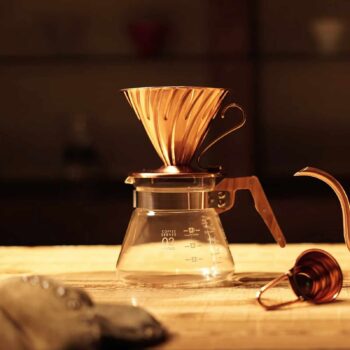 Ensemble de préparation de café filtre Hario V60 en cuivre, comprenant un dripper conique et une carafe en verre graduée, sur une table en bois.