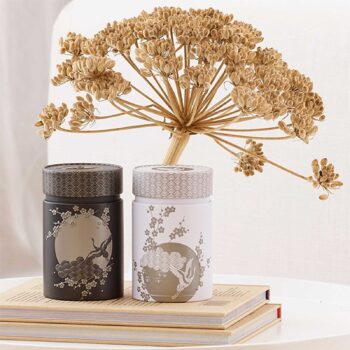 Deux boîtes à thé Eigenart Komorebi, une anthracite et une blanche, décorées de grues et de fleurs de cerisier, sur des livres