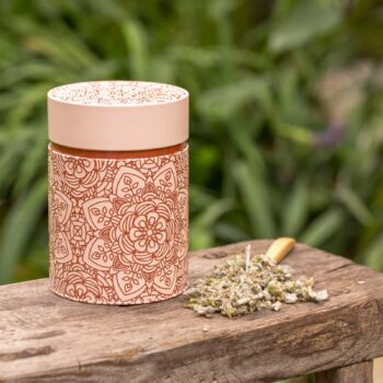 Boîte à thé Eigenart Mandala Powder rose poudré avec motifs embossés, à côté d'une cuillère en bois remplie d'herbes séchées sur un banc en bois