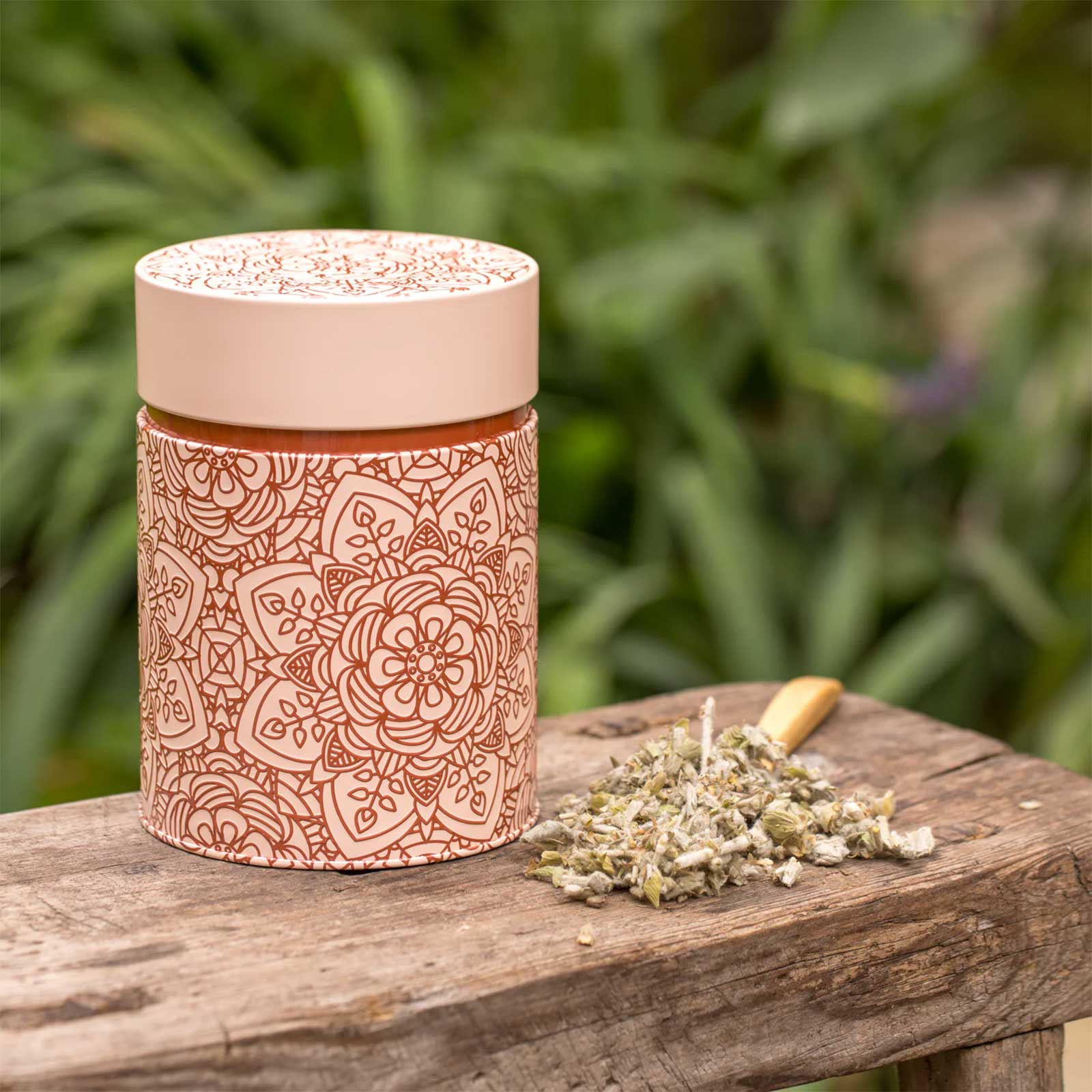 Boîte à thé Eigenart Mandala Powder rose poudré avec motifs embossés, à côté d'une cuillère en bois remplie d'herbes séchées sur un banc en bois